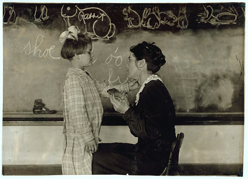 מוסדות מסוימים התעקשו ללמד ילדים עם לקויות שמיעה לדבר ולקרוא שפתיים. בתמונה: ילדה חירשת חשה את תנועות הפה והגרון בעת להגיית המילה "Fish" בבית ספר לחירשים  באוקלהומה, 1917 | מקור: Hine, Lewis Wickes; National Child Labor Committee Collection - Library of Congress Catalog