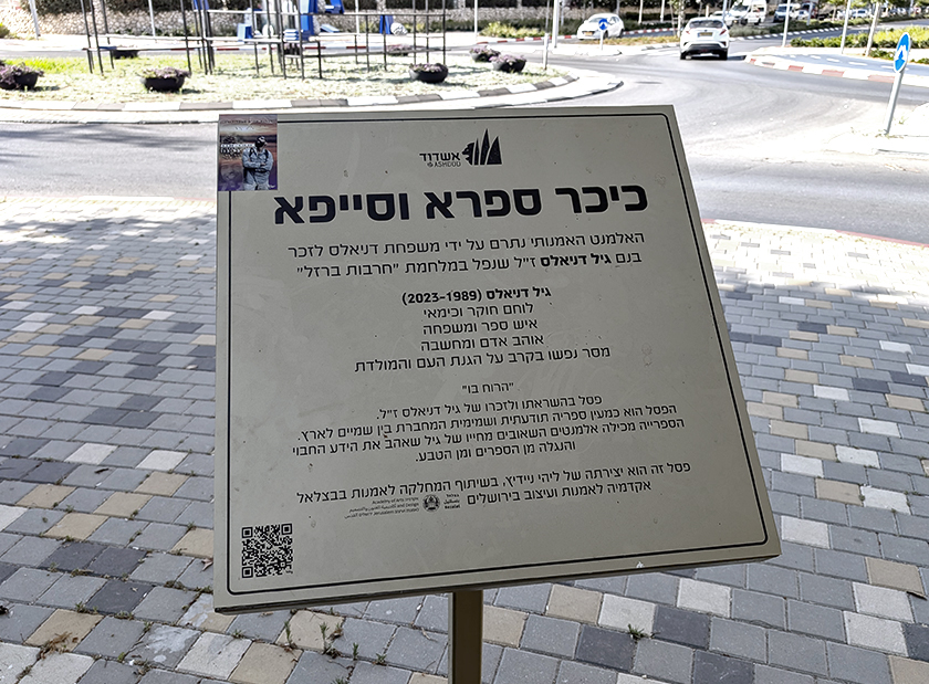 הנצחה פיזית ודיגיטלית. כיכר, פסל סביבתי ולוחית מידע עם קוד QR להנצחת סמל ראשון גיל דניאלס באשדוד | מקור: מקף־עברי, ויקימדיה