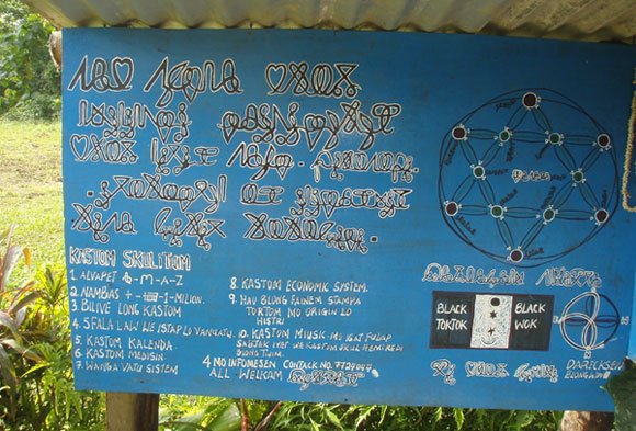 שלט הכניסה לבית הספר. שמו מופיע למעלה בכתב בוסטרופידוני: "המוסד המלנזי למדע, פילוסופיה, אנושיות וטכנולוגיה". מתחת מפורטים מקצועות הלימוד באנגלית משובשת | צילום: Tabisini, ויקיפדיה